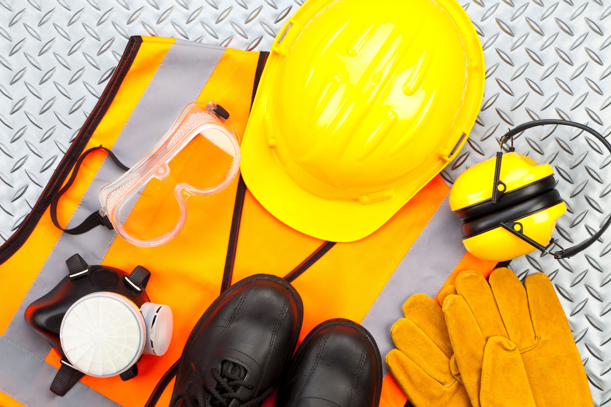 Um capacete amarelo, óculos de segurança, luvas de trabalho, botas de segurança, um colete refletivo e protetores auriculares dispostos sobre uma superfície texturizada.